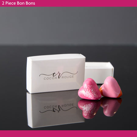 Two pink bon bons in front of a white box with 'Cocoa Rouge' branding on a reflective surface.