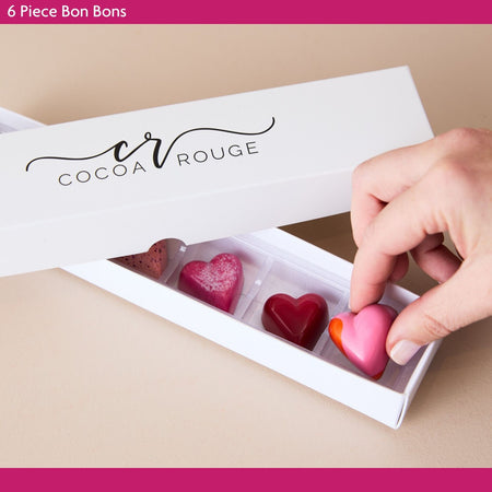 Cocoa Rouge chocolate bon  bons in a box with a hand reaching in to pick up a pink heart-shaped chocolate.