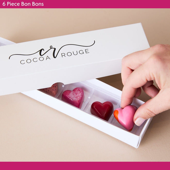 Cocoa Rouge chocolate bon  bons in a box with a hand reaching in to pick up a pink heart-shaped chocolate.