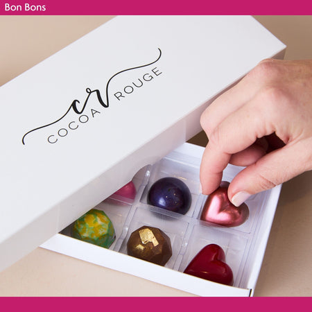 Hand reaching into a box of colorful chocolates labeled 'Cocoa Rouge'.