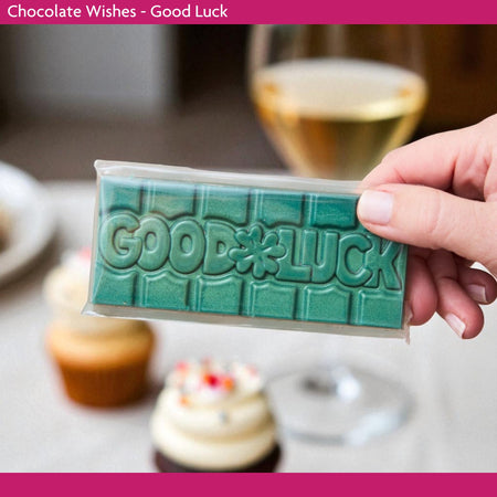 Hand holding a green 'Good Luck' chocolate bar with a blurred background of cupcakes and a glass.