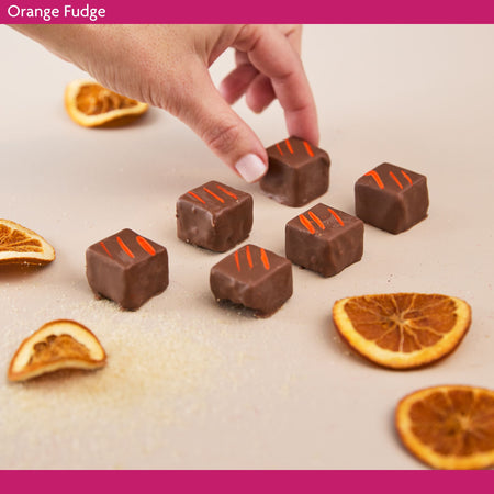 Chocolate fudge squares with orange zest on a surface with dried oranges, and a hand reaching towards one of the fudge pieces.