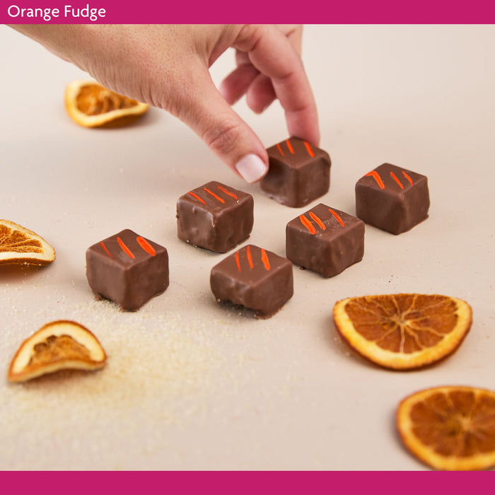 Chocolate fudge squares with orange zest on a surface with dried oranges, and a hand reaching towards one of the fudge pieces.
